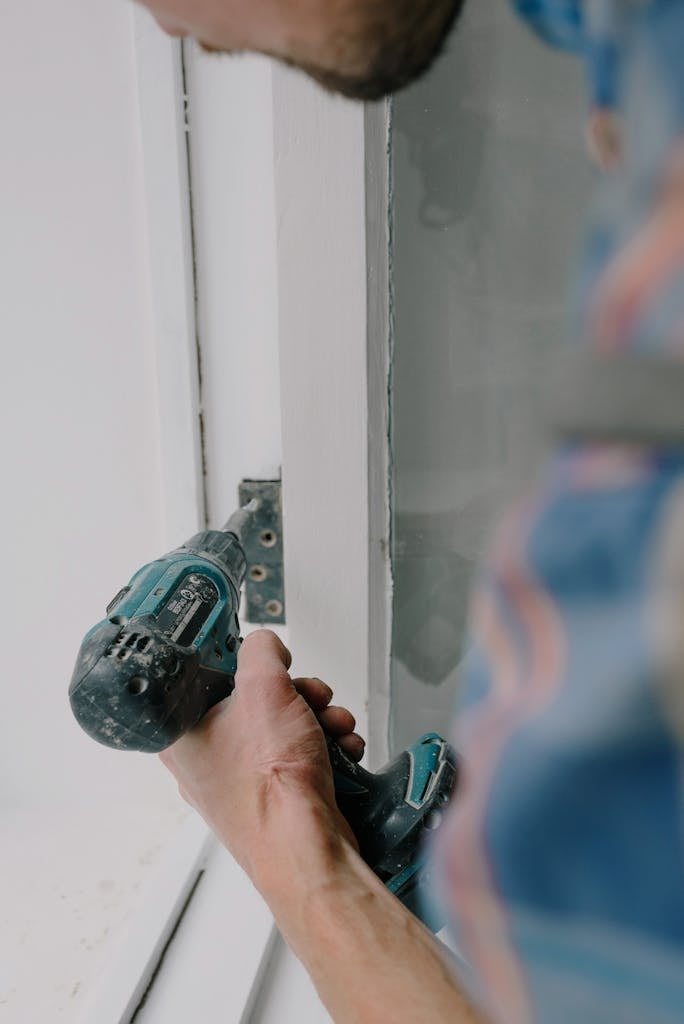 Crop bearded guy installing new wooden window with contemporary screwdriver and metal hinge during renovation process in apartment