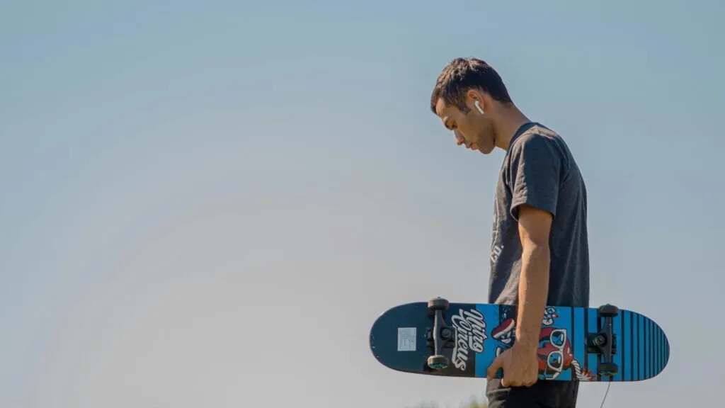 Teenager holding a skateboard, wearing AirPods, under clear blue sky.