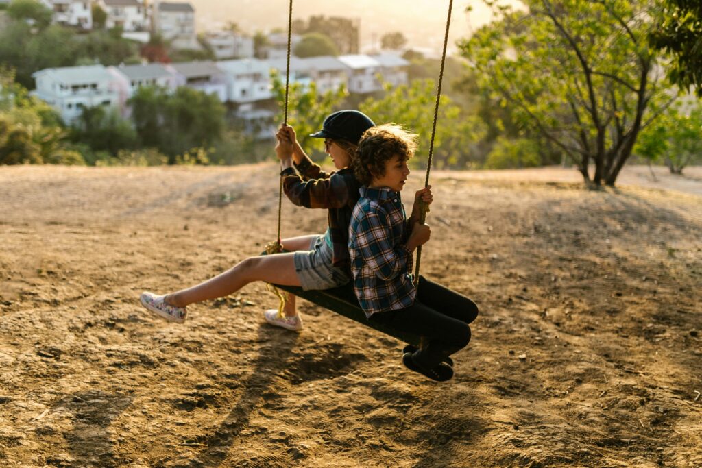 Two children enjoy a playful swing ride outdoors at sunset.