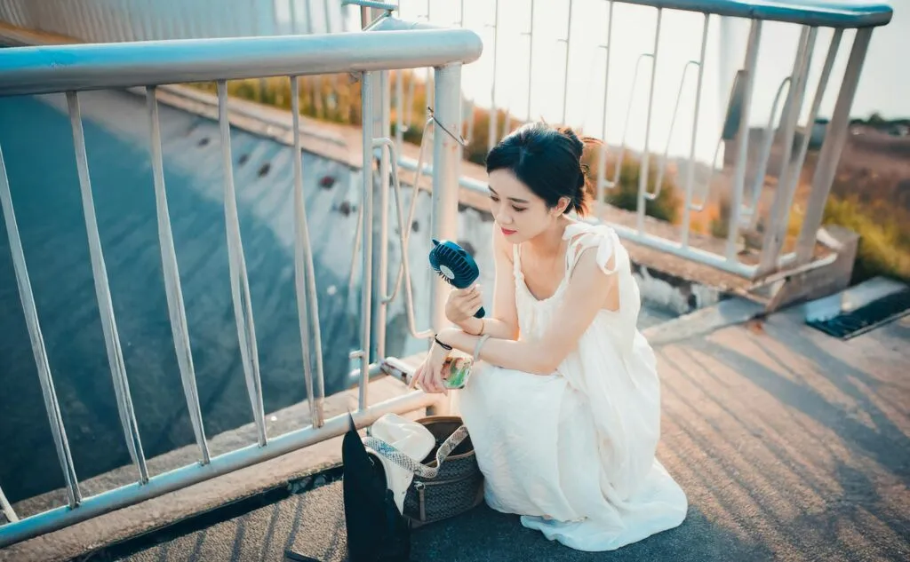 Woman in a white dress cools down with a portable fan on a sunny day outdoors.